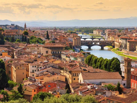 Vista panorámica de Florencia, Italia, mostrando el río Arno con varios puentes, incluido el famoso Ponte Vecchio, rodeado de edificios históricos y tejados rojizos bajo un cielo cálido al atardecer.
