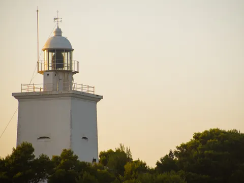 Faro blanco de Santa Pola, Alicante, destacando sobre un horizonte de color naranja pálido al amanecer, con árboles debajo.