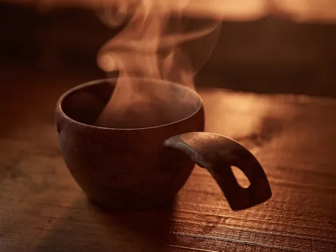 Taza rústica de madera con bebida caliente humeante sobre una mesa de madera iluminada por luz cálida.