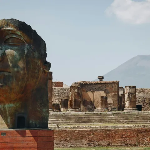 Cabeza de bronce en Pompeya con el Monte Vesubio y las ruinas de columnas romanas de fondo.