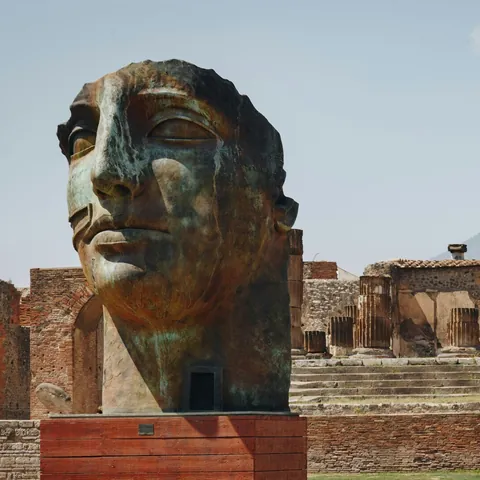 Cabeza de bronce en Pompeya con el Monte Vesubio y las ruinas de columnas romanas de fondo.