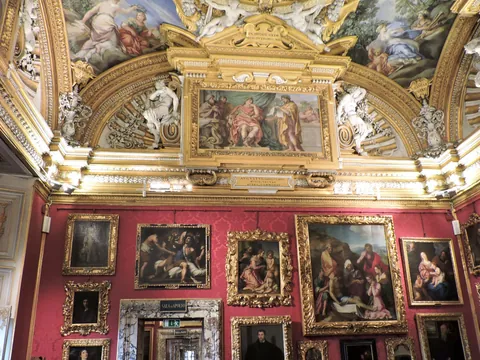 Sala del Palacio Pitti en Florencia con paredes rojas cubiertas de pinturas renacentistas en marcos dorados, techo decorado con frescos y molduras ornamentadas en oro.