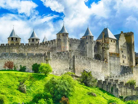 Vista de las murallas y torres medievales de Carcasona entre colinas verdes, bajo un cielo dramático.