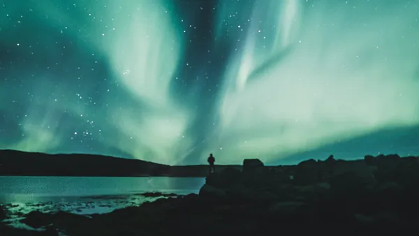 Aurora boreal iluminando el cielo nocturno de Islandia, con una silueta humana observando junto a un lago.