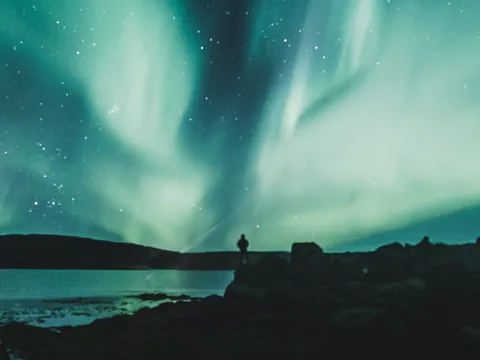 Aurora boreal iluminando el cielo nocturno de Islandia, con una silueta humana observando junto a un lago.