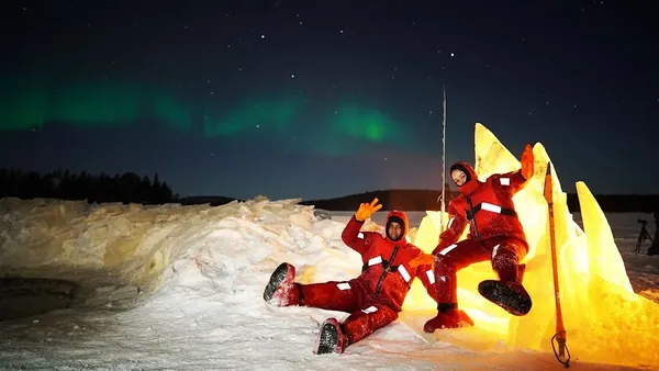 Dos personas con trajes térmicos rojos sentadas sobre nieve iluminada por luz cálida, bajo un cielo nocturno con aurora boreal verde en un paisaje ártico.