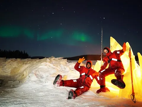 Dos personas con trajes térmicos rojos sentadas sobre nieve iluminada por luz cálida, bajo un cielo nocturno con aurora boreal verde en un paisaje ártico.