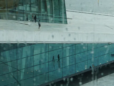 Reflejo en los cristales de la Ópera de Oslo, Noruega, con techo de mármol blanco y personas paseando bajo la lluvia. (