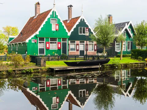 Casas típicas holandesas de madera verde con tejados a dos aguas, situadas junto a un canal en el que se reflejan, rodeadas de árboles y vegetación en Zaanse Schans, Países Bajos.