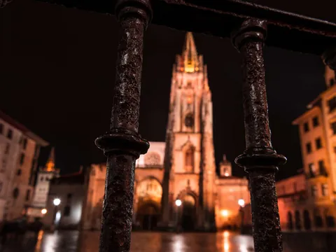 Vista nocturna de la Catedral de San Salvador de Oviedo a través de una barandilla, con la torre gótica iluminada en el centro.