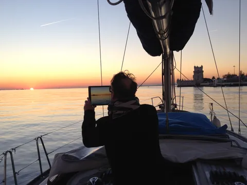 Persona sentada en la cubierta de un velero durante la puesta de sol sobre el río Tajo en Lisboa, con el cielo iluminado en tonos cálidos.