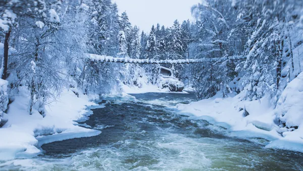 Río con corriente rodeado de bosque nevado, con un puente colgante cubierto de nieve y una cabaña al fondo.
