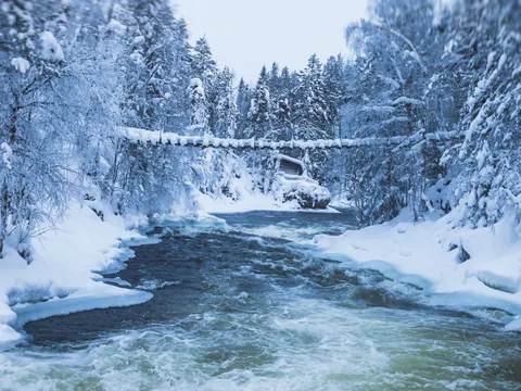 Río con corriente rodeado de bosque nevado, con un puente colgante cubierto de nieve y una cabaña al fondo.