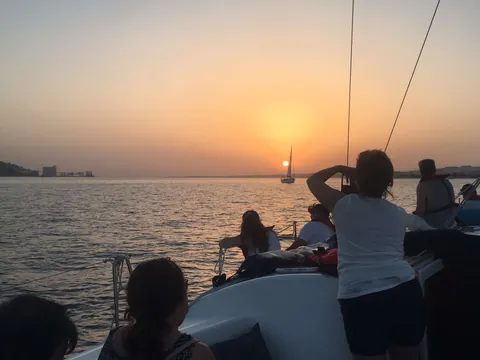 Grupo de personas disfrutando de un paseo en velero durante la puesta de sol sobre el río Tajo en Lisboa, con el cielo iluminado en tonos cálidos y reflejos en el agua.