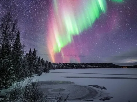 Aurora boreal con tonos verdes y rosados iluminando el cielo nocturno sobre un lago congelado y bosque nevado en un paisaje ártico.