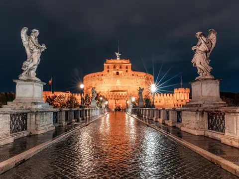Castillo Sant'Angelo en Roma iluminado de noche, visto desde el Puente Sant'Angelo con estatuas barrocas y reflejos en el pavimento mojado.