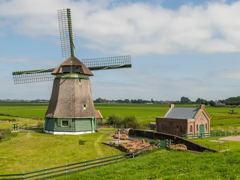 Molino de viento tradicional neerlandés junto a una pequeña casa de ladrillo, rodeados de campos verdes y canales.