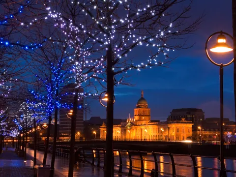 Vista nocturna del edificio histórico Custom House iluminado en Dublín, con árboles decorados con luces azules y blancas junto al río y farolas encendidas en el paseo.
