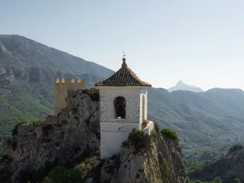 Vista aérea del campanario de Guadalest sobre un peñasco rocoso con montañas y vegetación de fondo.
