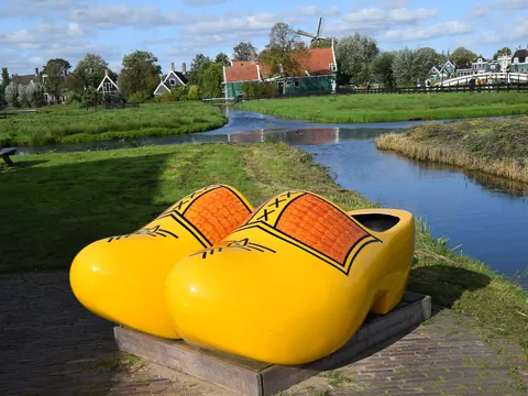 Zuecos de madera gigantes amarillos junto a un canal, con casas tradicionales neerlandesas y molinos de fondo en un paisaje típico de Ámsterdam.