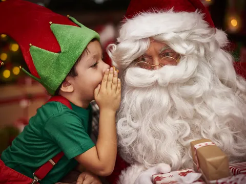 Niño vestido de duende navideño susurrando al oído de Papá Noel, rodeados de regalos en un ambiente festivo típico de Navidad.